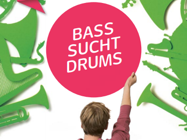 "Bass sucht Drums" Schriftzug wird gehalten von jungen Mann, er steht abgewendet mit dem Rücken zu uns